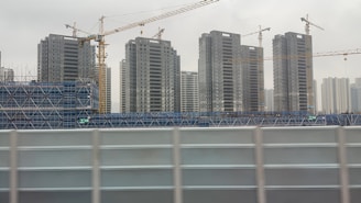 Several tall apartment buildings are under construction, surrounded by blue scaffolding and cranes. The sky appears overcast, contributing to a somewhat dreary atmosphere.