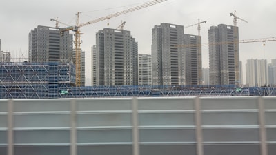Several tall apartment buildings are under construction, surrounded by blue scaffolding and cranes. The sky appears overcast, contributing to a somewhat dreary atmosphere.