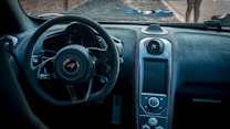 The image features the interior of a sports car, highlighting a steering wheel with a distinctive logo in the center. The dashboard is designed with a sleek, modern aesthetic, showcasing a variety of controls and a large display screen. The materials appear to be high-end, possibly leather and carbon fiber, contributing to a luxurious feel. Outside the windshield, a blurred view of a street and partially visible figures can be seen.