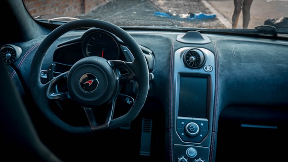 The image features the interior of a sports car, highlighting a steering wheel with a distinctive logo in the center. The dashboard is designed with a sleek, modern aesthetic, showcasing a variety of controls and a large display screen. The materials appear to be high-end, possibly leather and carbon fiber, contributing to a luxurious feel. Outside the windshield, a blurred view of a street and partially visible figures can be seen.