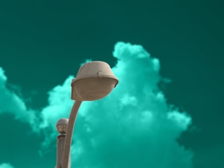 A streetlight with a futuristic design stands against a striking teal-green sky, with soft, fluffy clouds adding depth to the background. The light fixture is off, and a small satellite dish is visible on the pole.