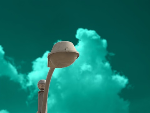 A streetlight with a futuristic design stands against a striking teal-green sky, with soft, fluffy clouds adding depth to the background. The light fixture is off, and a small satellite dish is visible on the pole.