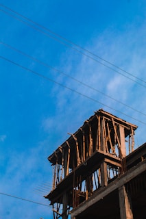 A partially constructed building with wooden scaffolding is set against a bright blue sky. Electrical wires are visible, stretching diagonally across the scene. The structure appears to be in the early stages of development, with exposed beams and supports.