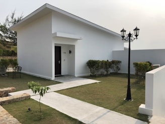 A minimalist white house with a slanted roof and a black door marked with the number 1706. It is surrounded by a well-maintained green lawn and hedges. A black vintage-style street lamp stands near a paved pathway leading to the entrance. There is a chair placed beside the wall in the backyard.