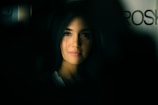 A portrait of a person with long, dark hair, softly illuminated against a dark background. The lighting emphasizes the gentle features of the person's face, creating a contrast between light and shadow. The background is blurred, adding an element of depth.
