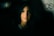 A portrait of a person with long, dark hair, softly illuminated against a dark background. The lighting emphasizes the gentle features of the person's face, creating a contrast between light and shadow. The background is blurred, adding an element of depth.