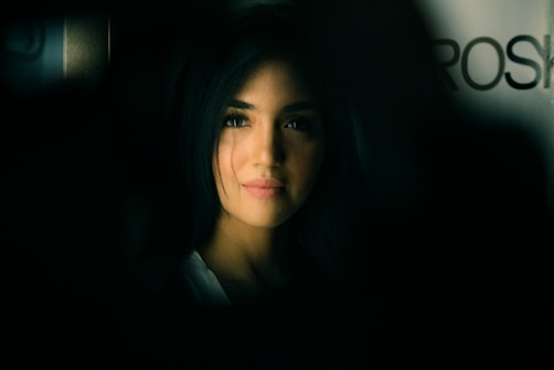 A portrait of a person with long, dark hair, softly illuminated against a dark background. The lighting emphasizes the gentle features of the person's face, creating a contrast between light and shadow. The background is blurred, adding an element of depth.
