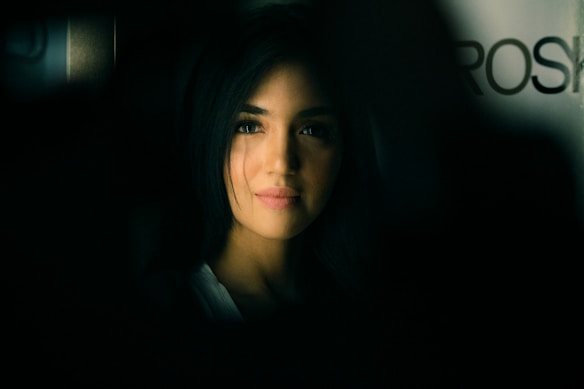 A portrait of a person with long, dark hair, softly illuminated against a dark background. The lighting emphasizes the gentle features of the person's face, creating a contrast between light and shadow. The background is blurred, adding an element of depth.