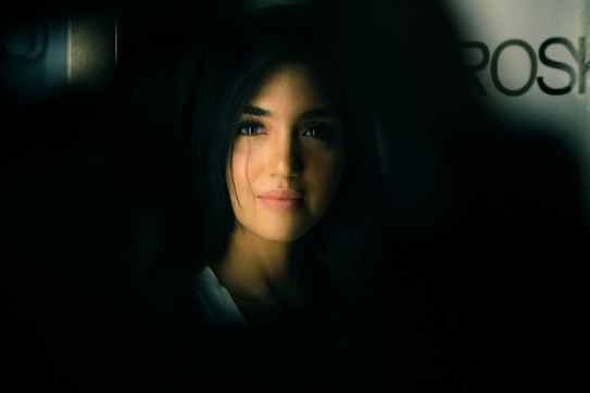 A portrait of a person with long, dark hair, softly illuminated against a dark background. The lighting emphasizes the gentle features of the person's face, creating a contrast between light and shadow. The background is blurred, adding an element of depth.