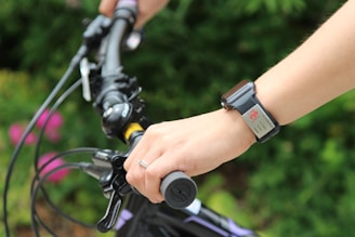 A person holding the handlebars of a bicycle, wearing a smartwatch on the left wrist. The background is blurred with greenery, suggesting an outdoor setting. The focus is on the hand and the watch, which displays a red icon.