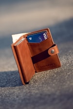 A brown leather wallet standing upright on a textured gray surface. It is open, revealing a blue Visa credit card and some cash tucked inside. The wallet's brand name 'KALIBRADO' is embossed on the leather.