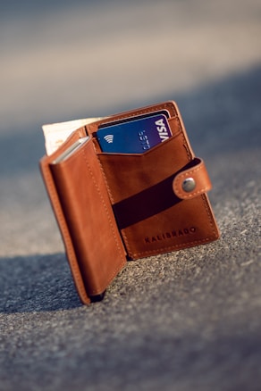 A brown leather wallet standing upright on a textured gray surface. It is open, revealing a blue Visa credit card and some cash tucked inside. The wallet's brand name 'KALIBRADO' is embossed on the leather.