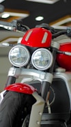 A close-up view of a motorcycle featuring dual round headlights, a prominent red fuel tank, and a sturdy front suspension. The motorcycle has a modern and robust design with a focus on the front wheel and tire. The showroom setting provides a sleek and polished look.