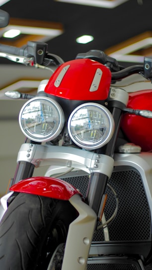 A close-up view of a motorcycle featuring dual round headlights, a prominent red fuel tank, and a sturdy front suspension. The motorcycle has a modern and robust design with a focus on the front wheel and tire. The showroom setting provides a sleek and polished look.