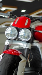 A close-up view of a motorcycle featuring dual round headlights, a prominent red fuel tank, and a sturdy front suspension. The motorcycle has a modern and robust design with a focus on the front wheel and tire. The showroom setting provides a sleek and polished look.