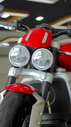 A close-up view of a motorcycle featuring dual round headlights, a prominent red fuel tank, and a sturdy front suspension. The motorcycle has a modern and robust design with a focus on the front wheel and tire. The showroom setting provides a sleek and polished look.
