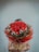 A person is holding a large bouquet of red roses adorned with small white flowers and green leaves. The bouquet is wrapped in brown and red paper with a decorative ribbon featuring the word 'love.' The background is a plain, light-colored wall.
