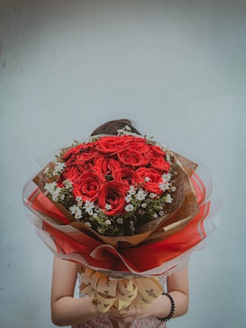 A person is holding a large bouquet of red roses adorned with small white flowers and green leaves. The bouquet is wrapped in brown and red paper with a decorative ribbon featuring the word 'love.' The background is a plain, light-colored wall.