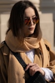 A woman with shoulder-length dark hair is wearing round sunglasses with a reflective surface. She is dressed in a beige coat over a white shirt and wrapped in a light brown scarf. Her hand is holding a textured black handbag strap.