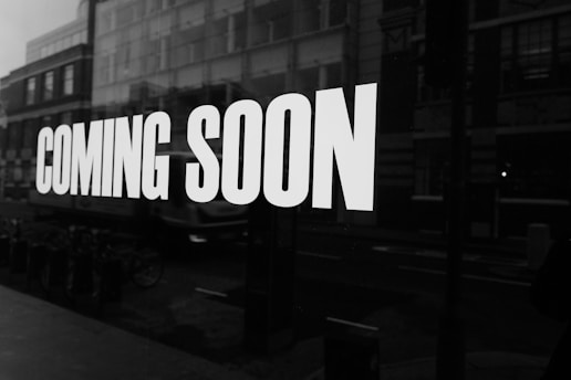 Large white text on a dark background reads 'COMING SOON,' set against the reflective surface of a window that reveals a blurred view of urban buildings and parked bicycles outside.