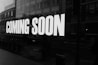 Large white text on a dark background reads 'COMING SOON,' set against the reflective surface of a window that reveals a blurred view of urban buildings and parked bicycles outside.
