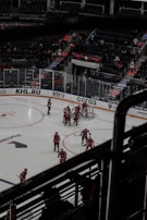 A group of ice hockey players in red uniforms are gathered around a goal on an indoor rink. The arena has empty seats with a few spectators, and there is dim lighting emphasizing the dramatic atmosphere of the match.