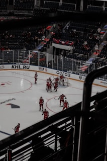 A group of ice hockey players in red uniforms are gathered around a goal on an indoor rink. The arena has empty seats with a few spectators, and there is dim lighting emphasizing the dramatic atmosphere of the match.
