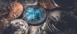 A close-up view of various coins, including pennies and quarters, with a focus on one coin that appears worn and has a distinctive blue-green patina. The lighting accentuates the metallic textures and intricate designs on the coins.