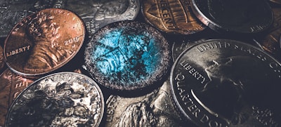 A close-up view of various coins, including pennies and quarters, with a focus on one coin that appears worn and has a distinctive blue-green patina. The lighting accentuates the metallic textures and intricate designs on the coins.