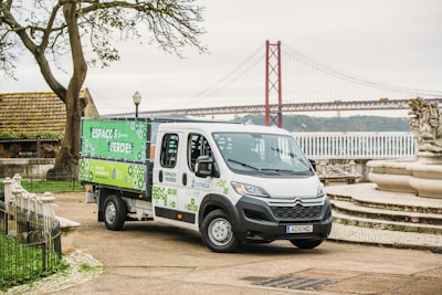 A utility vehicle with a green and white design is parked on a paved area next to a decorative stone fountain and large trees. In the background, a red suspension bridge stretches across a wide river, and a cloudy sky adds to the scene. The vehicle bears logos and text related to green spaces.