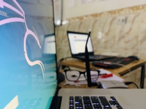 A close-up of an office or home workspace with a focus on two laptops. One laptop displays an operating system with a graphical interface, possibly Linux-based, showing a stylized dragon logo. Another laptop is open and appears to be used for browsing, with some blurred white and red colors on the screen. In the foreground, a pair of glasses rests on the table, along with several papers and a plastic card. The background has a tiled wall, adding a casual or homely feel to the workspace.