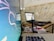 A close-up of an office or home workspace with a focus on two laptops. One laptop displays an operating system with a graphical interface, possibly Linux-based, showing a stylized dragon logo. Another laptop is open and appears to be used for browsing, with some blurred white and red colors on the screen. In the foreground, a pair of glasses rests on the table, along with several papers and a plastic card. The background has a tiled wall, adding a casual or homely feel to the workspace.