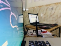 A close-up of an office or home workspace with a focus on two laptops. One laptop displays an operating system with a graphical interface, possibly Linux-based, showing a stylized dragon logo. Another laptop is open and appears to be used for browsing, with some blurred white and red colors on the screen. In the foreground, a pair of glasses rests on the table, along with several papers and a plastic card. The background has a tiled wall, adding a casual or homely feel to the workspace.