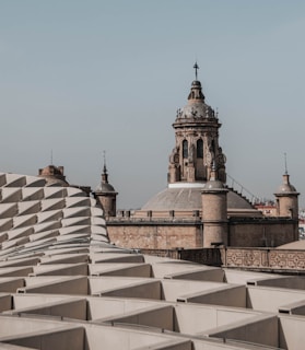 An architectural scene featuring a historic building with a large, ornate dome and spire, surrounded by smaller spires. In the foreground, there is a modern structure with a repeating geometric pattern of white panels.