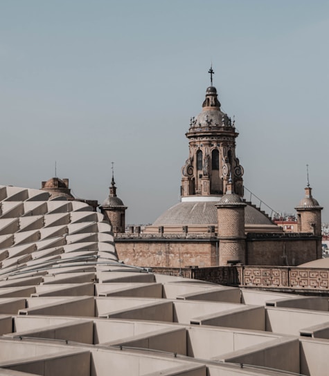 An architectural scene featuring a historic building with a large, ornate dome and spire, surrounded by smaller spires. In the foreground, there is a modern structure with a repeating geometric pattern of white panels.