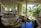 A spacious and well-lit indoor area featuring multiple levels connected by stairs, with a garden-like setting including a water feature, rocks, and lush plants. The area is open to natural light from large windows and columns, with tropical foliage visible outside.