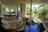 A spacious and well-lit indoor area featuring multiple levels connected by stairs, with a garden-like setting including a water feature, rocks, and lush plants. The area is open to natural light from large windows and columns, with tropical foliage visible outside.