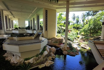 A spacious and well-lit indoor area featuring multiple levels connected by stairs, with a garden-like setting including a water feature, rocks, and lush plants. The area is open to natural light from large windows and columns, with tropical foliage visible outside.