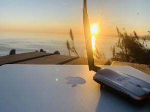 A sunset over the ocean is visible in the background, casting a warm glow. In the foreground, a laptop with a recognizable logo is placed on a wooden surface, next to a wireless antenna device. Tall grasses are silhouetted against the sky, adding a natural touch to the scene.