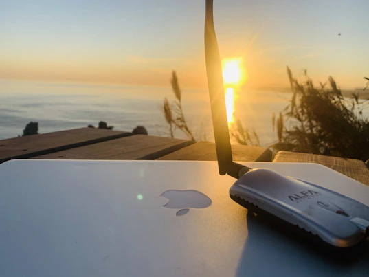 A sunset over the ocean is visible in the background, casting a warm glow. In the foreground, a laptop with a recognizable logo is placed on a wooden surface, next to a wireless antenna device. Tall grasses are silhouetted against the sky, adding a natural touch to the scene.