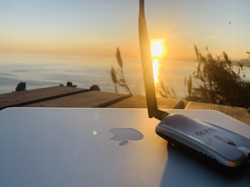 A sunset over the ocean is visible in the background, casting a warm glow. In the foreground, a laptop with a recognizable logo is placed on a wooden surface, next to a wireless antenna device. Tall grasses are silhouetted against the sky, adding a natural touch to the scene.