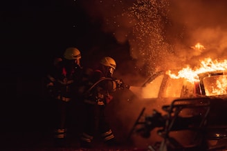 Two firefighters in protective gear are using a hose to extinguish a car engulfed in flames. Sparks and smoke fill the air, highlighting the intensity of the fire.