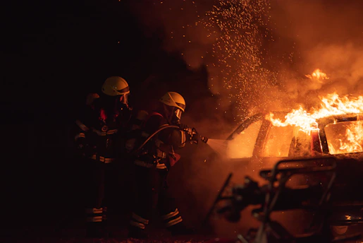 Two firefighters in protective gear are using a hose to extinguish a car engulfed in flames. Sparks and smoke fill the air, highlighting the intensity of the fire.