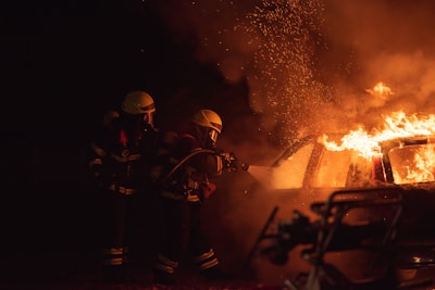 Two firefighters in protective gear are using a hose to extinguish a car engulfed in flames. Sparks and smoke fill the air, highlighting the intensity of the fire.