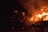 Two firefighters in protective gear are using a hose to extinguish a car engulfed in flames. Sparks and smoke fill the air, highlighting the intensity of the fire.