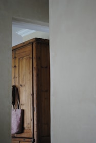 A wooden wardrobe partially visible through an open doorway in a softly lit room. The wardrobe is made of dark wood with visible grain, and a pinkish tote bag hangs from its edge. The light gray walls and soft shadows create a calm atmosphere.