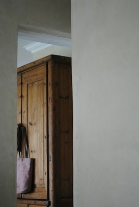 A wooden wardrobe partially visible through an open doorway in a softly lit room. The wardrobe is made of dark wood with visible grain, and a pinkish tote bag hangs from its edge. The light gray walls and soft shadows create a calm atmosphere.