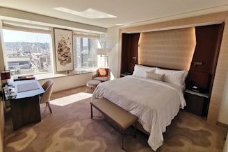 A modern, well-lit hotel room with a large bed featuring white linens and a cushioned headboard. There is a soft, light brown carpet on the floor and a bench at the foot of the bed. Large windows provide a view of a cityscape, allowing natural light to fill the room. To the left is a desk with a chair and some items placed on the surface, including water bottles. A comfortable chair and a small table are positioned near the windows. A piece of art hangs between the windows, and subtle, warm lighting enhances the cozy atmosphere.