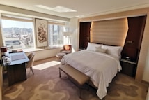 A modern, well-lit hotel room with a large bed featuring white linens and a cushioned headboard. There is a soft, light brown carpet on the floor and a bench at the foot of the bed. Large windows provide a view of a cityscape, allowing natural light to fill the room. To the left is a desk with a chair and some items placed on the surface, including water bottles. A comfortable chair and a small table are positioned near the windows. A piece of art hangs between the windows, and subtle, warm lighting enhances the cozy atmosphere.