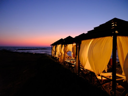 A series of cabanas with flowing white curtains are illuminated by a warm yellow light. They are positioned along a coastline during a vibrant and colorful sunset. The sky transitions from deep blue to orange and pink hues, reflecting over the calm sea.